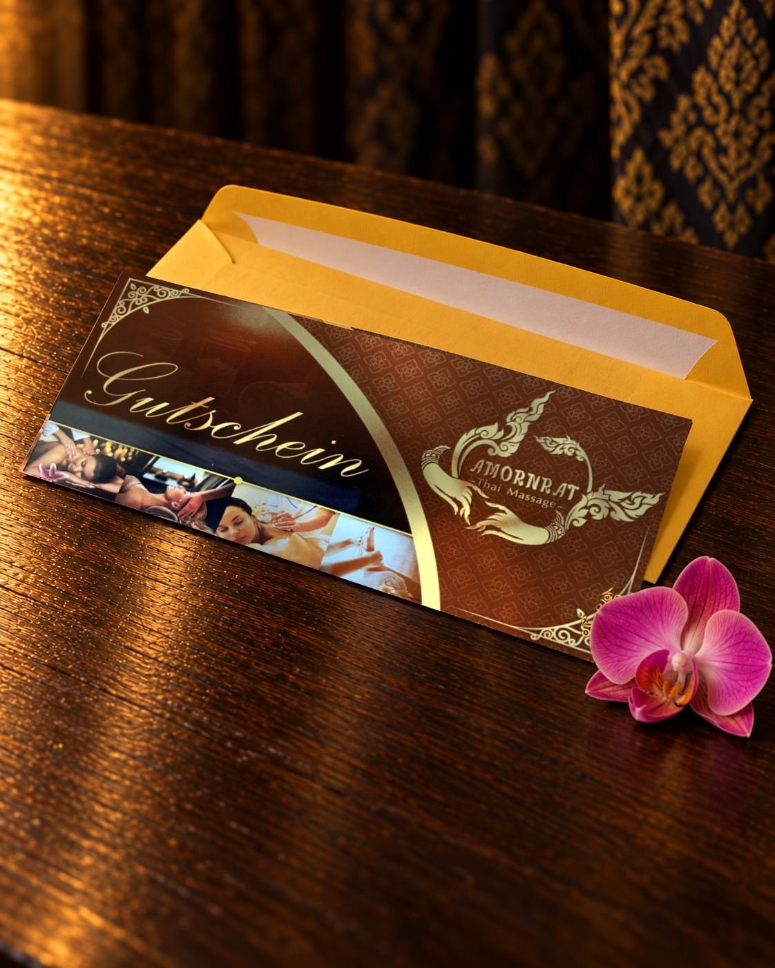 Geschenkgutschein von Amornrat Thai Massage: braune Karte mit goldener Schrift „Gutschein“ und dem Amornrat-Logo vor einem senfgelben Kuvert auf einem dunklen Holztisch, daneben eine pinke Orchidee